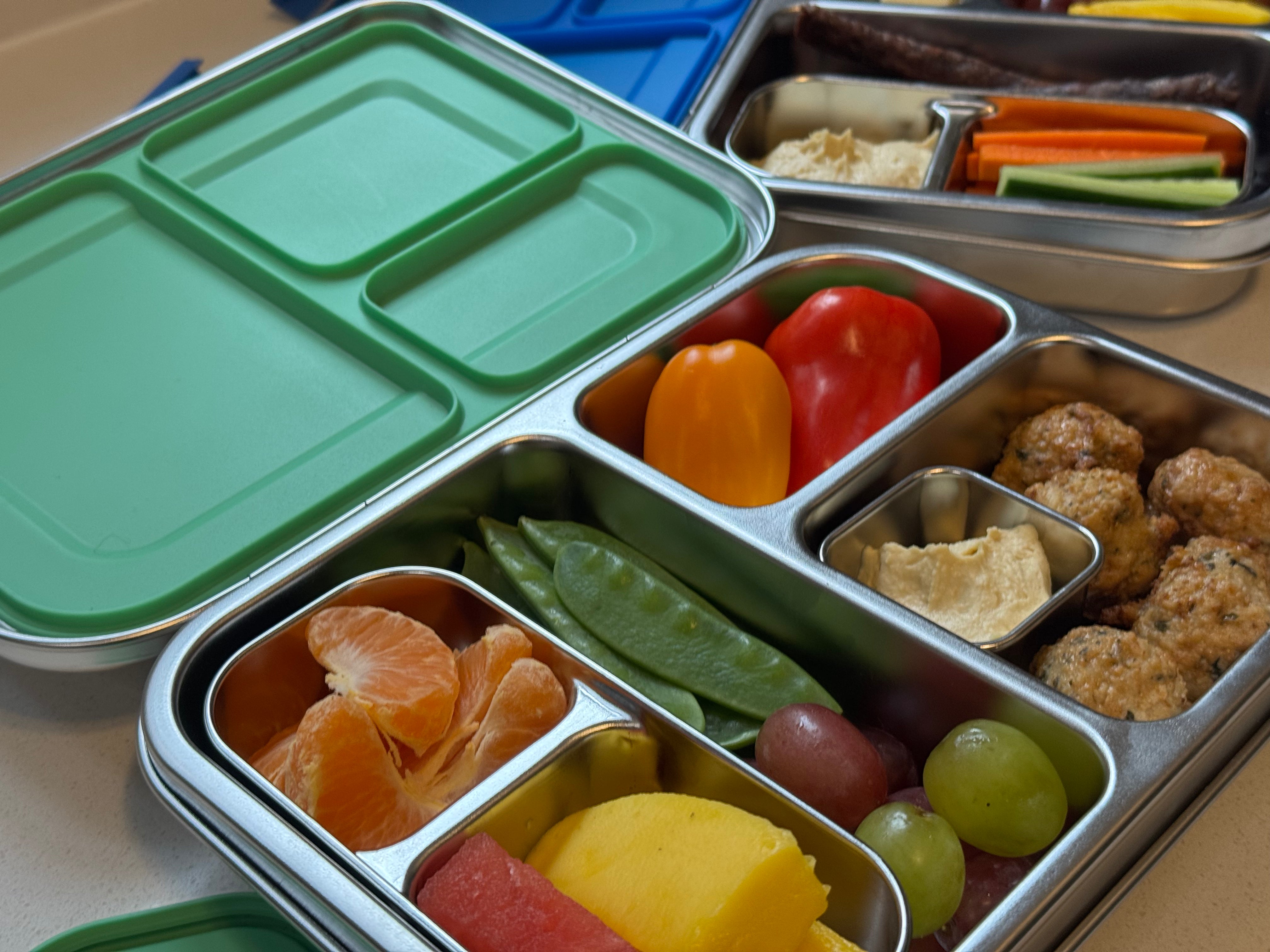 Stainless steel Finibini Bento lunchbox with compartments filled with various snacks including fruits and vegetables.